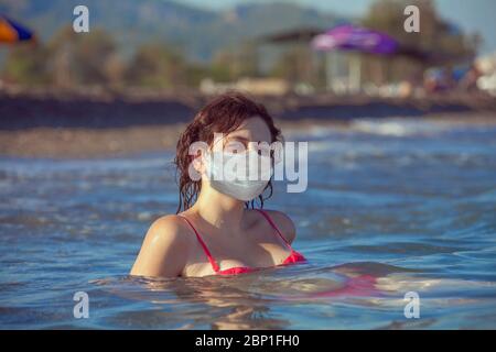 Une jeune femme nage dans l'eau de la mer sur le centre de villégiature avec un masque médical facial jetable pour la protection respiratoire sur son visage pendant le coronavirus Banque D'Images