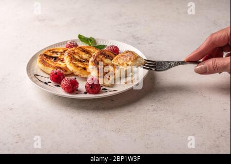 La main de la femme tient une fourchette avec un morceau de cheesecake sur une assiette de cheesecakes avec framboises et sirop. Mise au point sélective. Copier l'espace pour le texte Banque D'Images