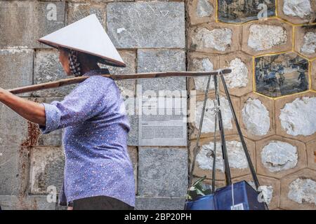 Hanoï, Vietnam - 1 mai 2018 : femme portant des paniers de fourche, portant un chapeau traditionnel, avec mur de pierre en arrière-plan Banque D'Images