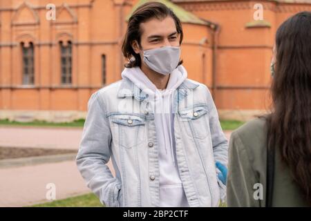 Un jeune homme heureux en veste en denim, sweat à capuche et masque de protection parlant à sa petite amie tout en se tenant dans un endroit public en milieu urbain Banque D'Images