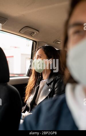 Jolie jeune femme brune en cuir veste et masque de protection écouter de la musique dans les écouteurs tout en rentant à la maison en taxi après le shopping Banque D'Images
