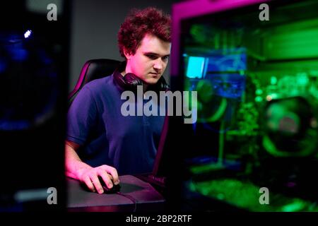 Un homme sérieux avec un casque autour du cou regardant l'écran d'ordinateur pendant le jeu cybersport en jouant dans un club contemporain Banque D'Images