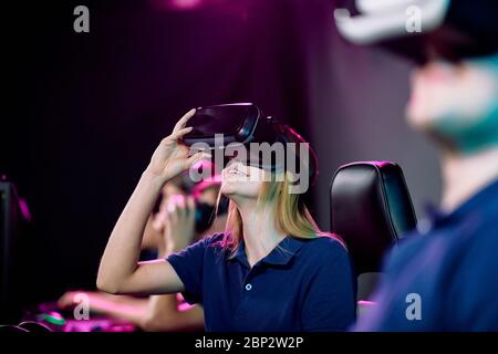 Une jeune gameuse vidéo heureuse dans un casque vr, assise parmi ses amis dans un club de jeux de cybersports et jouant en réseau Banque D'Images
