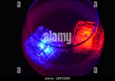 Les glaçons rouges et bleus brillants qui flottent dans une délicieuse boisson froide blanche dépolie sont placés dans un verre cognac sur un fond noir dépoli. Banque D'Images
