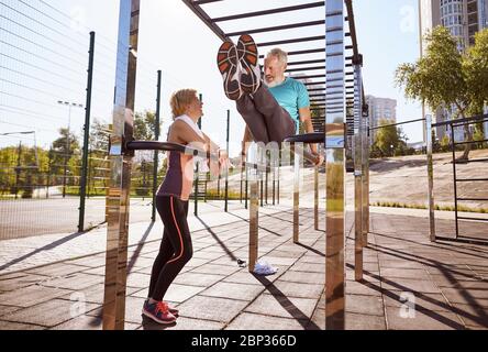 Sportif homme mature dans une grande forme faisant des repoussez sur des barres parallèles et parlant avec sa femme. Couple mature actif s'exerçant ensemble le matin Banque D'Images