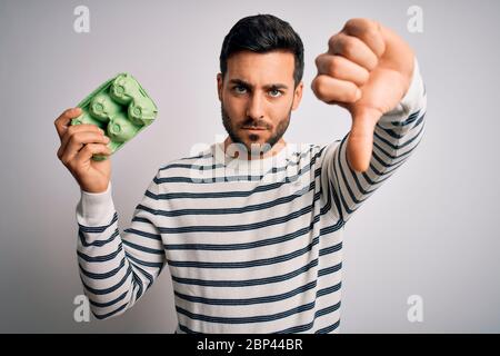 Jeune homme beau avec barbe tenant une boîte de carton d'œufs frais sur fond blanc avec visage en colère, signe négatif montrant le déplaît avec les pouces vers le bas, Banque D'Images