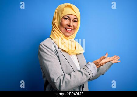 Femme d'affaires brunette d'âge moyen portant le hijab traditionnel musulman sur fond bleu invitant à entrer dans le sourire naturel avec la main ouverte Banque D'Images