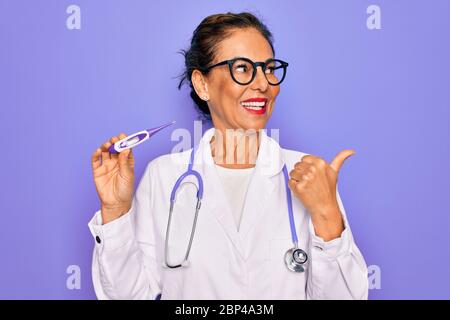 Femme médecin professionnelle d'âge moyen tenant un thermomètre médical pointant et montrant avec le pouce vers le côté avec le visage heureux souriant Banque D'Images