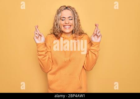 Jeune belle blonde sportive femme portant un sweat-shirt décontracté sur fond jaune gestante doigt croisé souriant avec espoir et les yeux fermés. Chance Banque D'Images