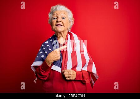 Senior magnifique femme patriotique aux cheveux gris portant le drapeau des états-unis sur fond rouge très heureux de pointer avec la main et le doigt Banque D'Images