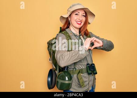 Jeune routard à tête rouge femme randonnée portant sac à dos et chapeau sur fond jaune souriant dans l'amour faisant le symbole de coeur forme avec les mains. Co romantique Banque D'Images