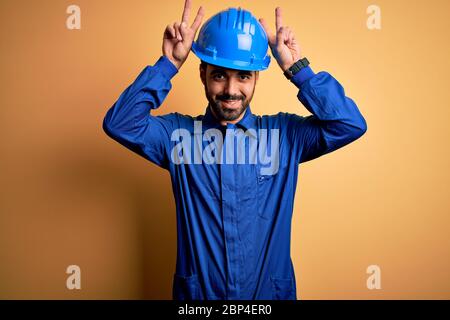 Mécanicien homme avec barbe portant l'uniforme bleu et casque de sécurité sur fond jaune posant drôle et fou avec les doigts sur la tête comme des oreilles de lapin, smili Banque D'Images