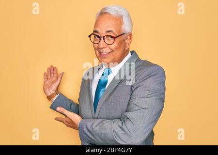 Homme d'affaires d'âge moyen à cheveux gris beau portant des lunettes sur fond jaune invitant à entrer dans le sourire naturel à main ouverte Banque D'Images