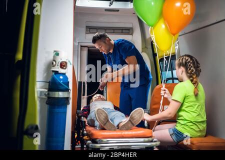 Homme dans un uniforme médical met un masque d'oxygène sur une fille allongé sur une civière, sa petite sœur regarde le travail du médecin Banque D'Images