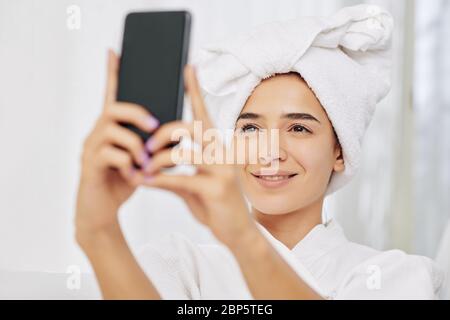 Jolie jeune femme en peignoir et serviette sur sa tête prenant le selfie Banque D'Images