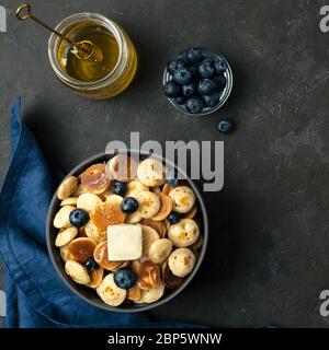 Plats tendance - crêpes, céréales, beurre et bleuets servis. Tas de mini crêpes aux céréales dans le boul sur fond sombre. Copier l'espace à droite pour le texte. Vue de dessus ou plan d'étapage. Recadrage carré Banque D'Images