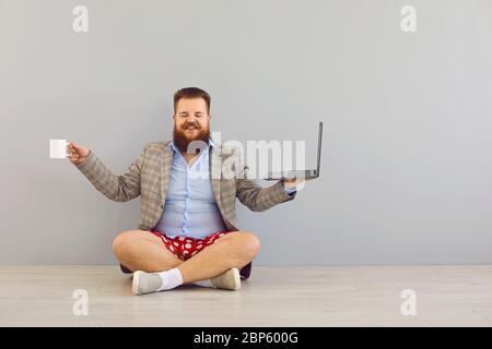 Concept de travail à distance en ligne indépendant. Drôle de barbu gros homme dans une veste et short rouge avec un ordinateur portable sur un fond gris. Banque D'Images