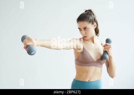 Mini portrait de la taille vers le haut de la jeune femme déterminée de lever des haltères tout en appréciant l'entraînement de fitness à la maison debout contre le mur blanc, l'espace de copie Banque D'Images