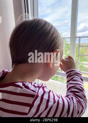 le garçon reste à la maison et regarde par la fenêtre. il manque ses amis et camarades de classe. Pendant la quarantaine, les enfants s'assoient à la maison en isolement. Banque D'Images