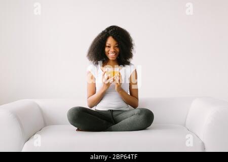 Une jeune femme afro-américaine assise dans la position lotus sur un lit blanc avec une tasse de thé Banque D'Images