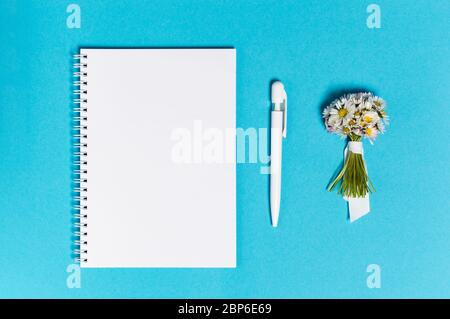 Carnet avec papier vierge, stylo et bouquet de fleurs de Marguerite sur fond bleu par le dessus. Freelance, rédaction ou planification de vacances. Posez à plat avec Banque D'Images
