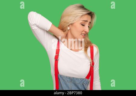 Dos douloureux et mal de cou. Portrait de femme blonde adulte épuisée en combinaison de denim sentant la tension dans les épaules et faisant le massage, la tension musculaire Banque D'Images