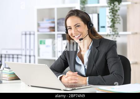Une femme de télévendeur heureuse avec un casque et un ordinateur portable se posant en regardant la caméra sur un bureau Banque D'Images
