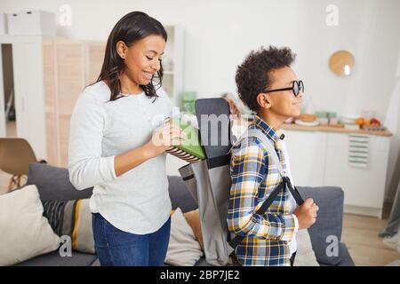 Portrait de la femme afro-américaine souriante qui emportait des livres dans un sac à dos tout en envoyant son fils à l'école Banque D'Images