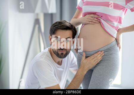 Jeune homme barbu mettant son oreille près de son abdomen enceinte Banque D'Images
