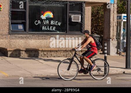 Montréal, CA - 17 mai 2020: Jeune homme avec masque pour la protection contre COVID-19 à vélo devant un arc-en-ciel dessinant sur la rue Masson. Banque D'Images