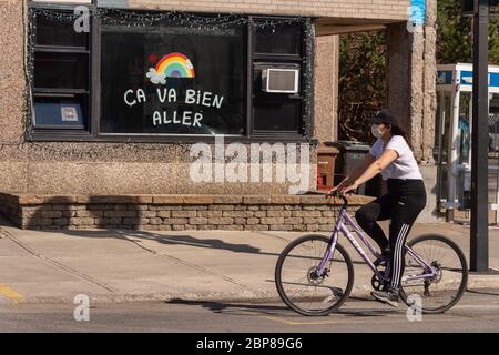 Montréal, CA - 17 mai 2020: Jeune femme avec masque pour la protection contre COVID-19 à vélo devant un arc-en-ciel dessinant sur la rue Masson. Banque D'Images