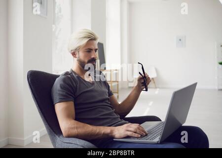 Travailler en ligne à domicile. Formation en ligne. Un jeune homme d'affaires vêque de façon décontractée travaille avec un ordinateur portable tout en étant assis dans un fauteuil dans une chambre résidentielle. Banque D'Images