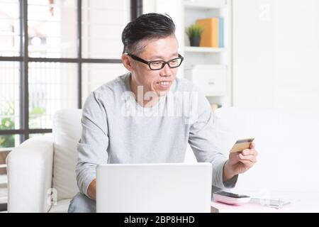 Portrait de l'homme asiatique mature des années 50 utilisant l'ordinateur faisant des achats en ligne avec carte de crédit, assis sur un canapé à la maison, retraité senior à l'intérieur vivant de la farce Banque D'Images