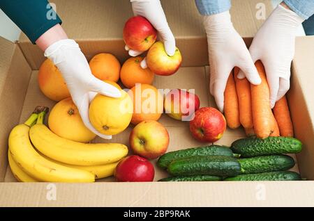 Les bénévoles en gants médicaux de protection mettent les fruits et les légumes dans la boîte de dons. Banque D'Images