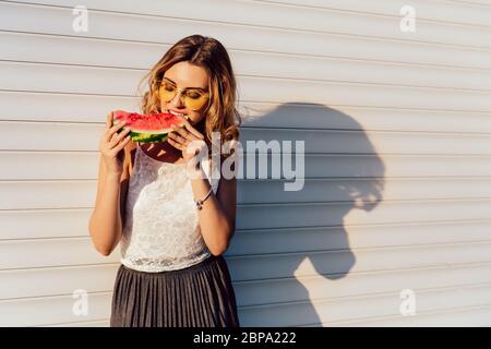 Charmante femme séduisante en mangeant une pastèque lunettes, debout contre le mur, à l'extérieur en milieu urbain. Vêtus de blouses et de jupes d'été. Banque D'Images