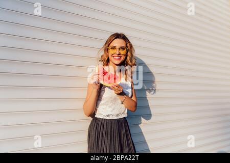 Happy Funny Girl en jaune lunettes détient un morceau de pastèque, debout près du mur urbain à l'extérieur. Habillé de vêtement élégant. Banque D'Images
