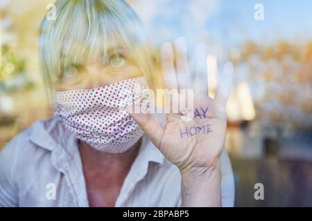 Une femme âgée avec masque tient la main avec un message de rester à la maison à la fenêtre pendant une pandémie de coronavirus Banque D'Images