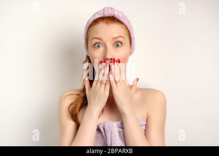 Mignonne fille à cheveux rouges se tient sur un fond blanc dans une serviette avec un bandage sur sa tête et dans surprise couvre la bouche avec les mains Banque D'Images