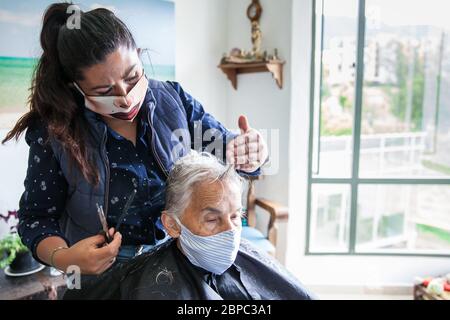 Femme âgée qui se coupe à la maison pendant la pandémie Covid-19 portant un masque facial Banque D'Images