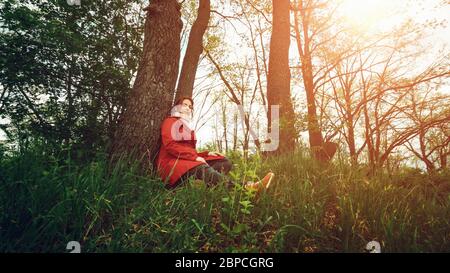 Jeune jolie femme en manteau rouge ayant le repos dans la forêt de printemps et regarde la caméra. Banque D'Images