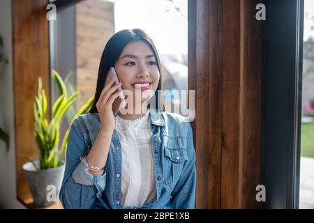 Femme aux cheveux sombres, penchée sur le cadre de la porte, souriante, parlant sur un téléphone portable Banque D'Images