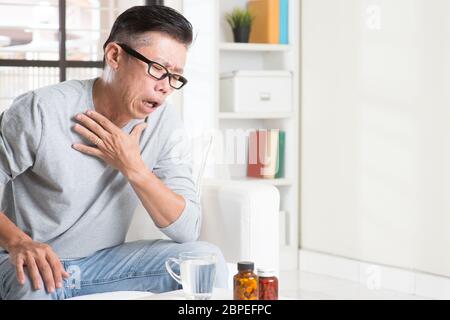 Portrait de 50s occasionnels mature Asian man toux, agissant sur poitrine avec expression douloureuse, assis sur le canapé à la maison, des médicaments et de l'eau sur la table. Banque D'Images
