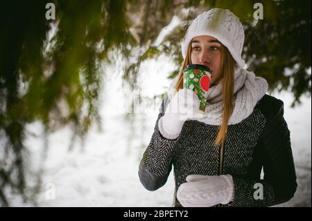 Portrait of cute femme en blanc foulard et un chapeau manteau en tricot sur l'extérieur de la neige fond et branches floues en hiver. girl est titulaire d'une tasse de café Banque D'Images