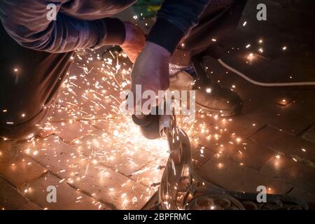 Vue avant des mains d'un homme travaillant sur une partie métallique d'un banc de jardin, utilisant une meuleuse électrique pendant que des étincelles volent dans le travail industriel Banque D'Images