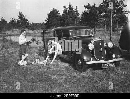 Vers les années 1940, à côté d'un lac dans une forêt ouverte, deux hommes, l'un debout sur l'herbe et l'autre assis sur un pied-à-pied ou un panneau de course d'une voiture de l'époque, une élégante berline quatre portes, ayant une tasse de thé, peut-être après un plongeon tôt le matin, Angleterre, Royaume-Uni. Banque D'Images