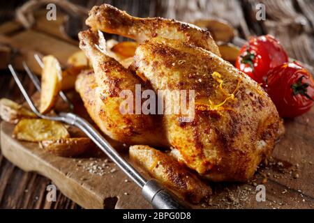 Jeune poulet grillé assaisonné de poussin, ou au printemps, de poulet servi entier sur une planche en bois rustique avec les quartiers de pommes de terre et de tomate Banque D'Images