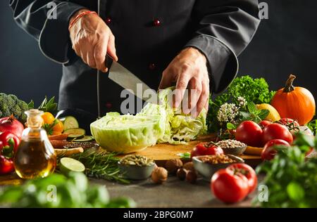 Chef chopping un chou frais sur une planche à découper en bois avec un couteau de cuisine, entourée d'un mélange de légumes pour le dîner Banque D'Images