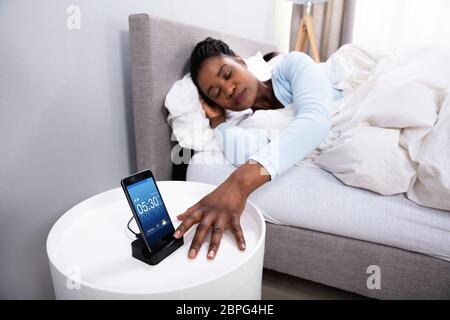 Jeune femme africaine près de couchage sur l'alarme du téléphone mobile dans la chambre Banque D'Images