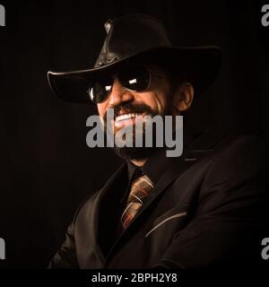 Portrait d'un beau homme élégant souriant en costume noir et chapeau de cow-boy noir sur fond noir. Banque D'Images