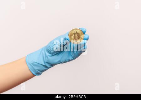 Vue latérale du profil gros plan de la main humaine dans des gants chirurgicaux bleus tenant et montrant le symbole bitcoin en main. Vue en intérieur, en studio, isolée sur le dos gris Banque D'Images